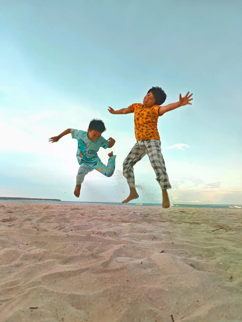 Two young boys jumping joyfully on a wide white-sand beach under a clear pale sky.