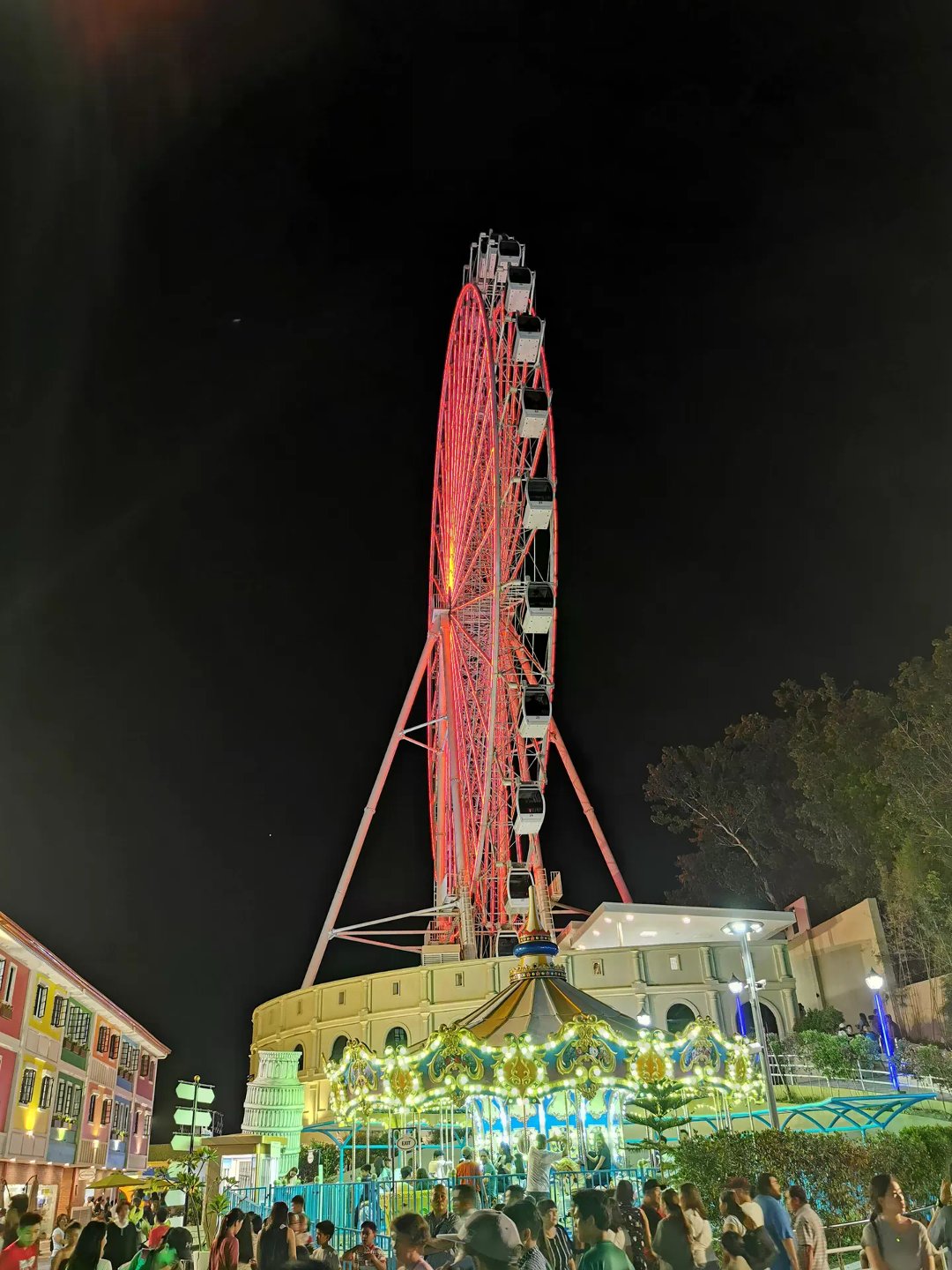 Brightly lit Ferris wheel and carousel at Anjo World Theme Park with crowds gathered at night.