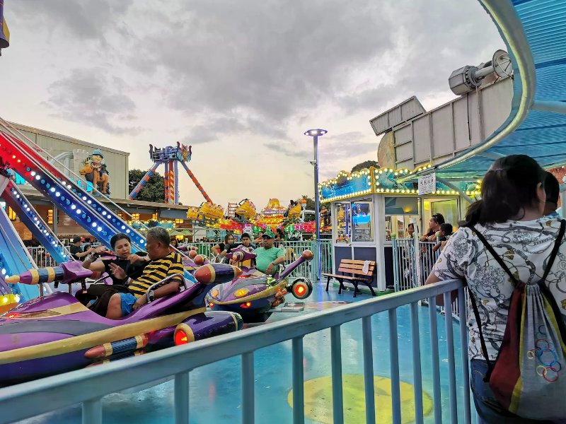 People riding purple rocket-shaped vehicles at Anjo World under a cloudy sunset sky.