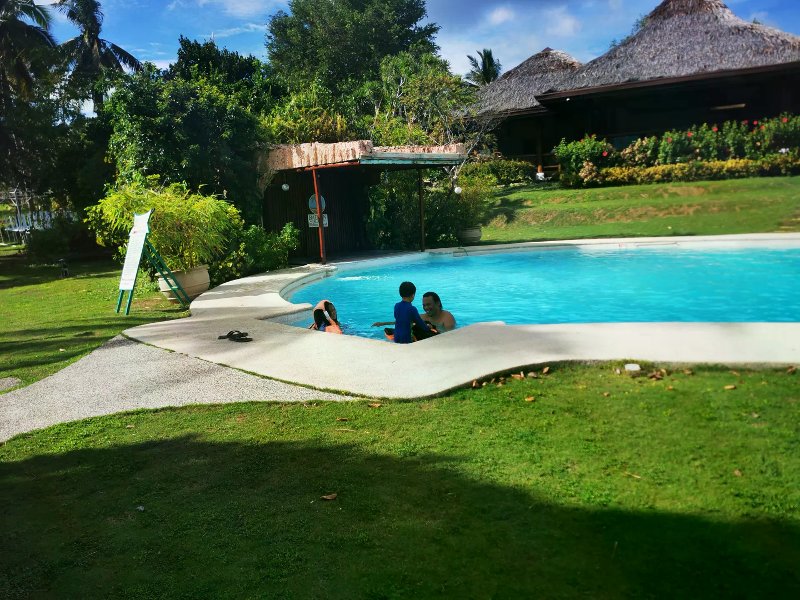 Family playing in a turquoise resort pool beside thatched-roof buildings and tropical gardens.