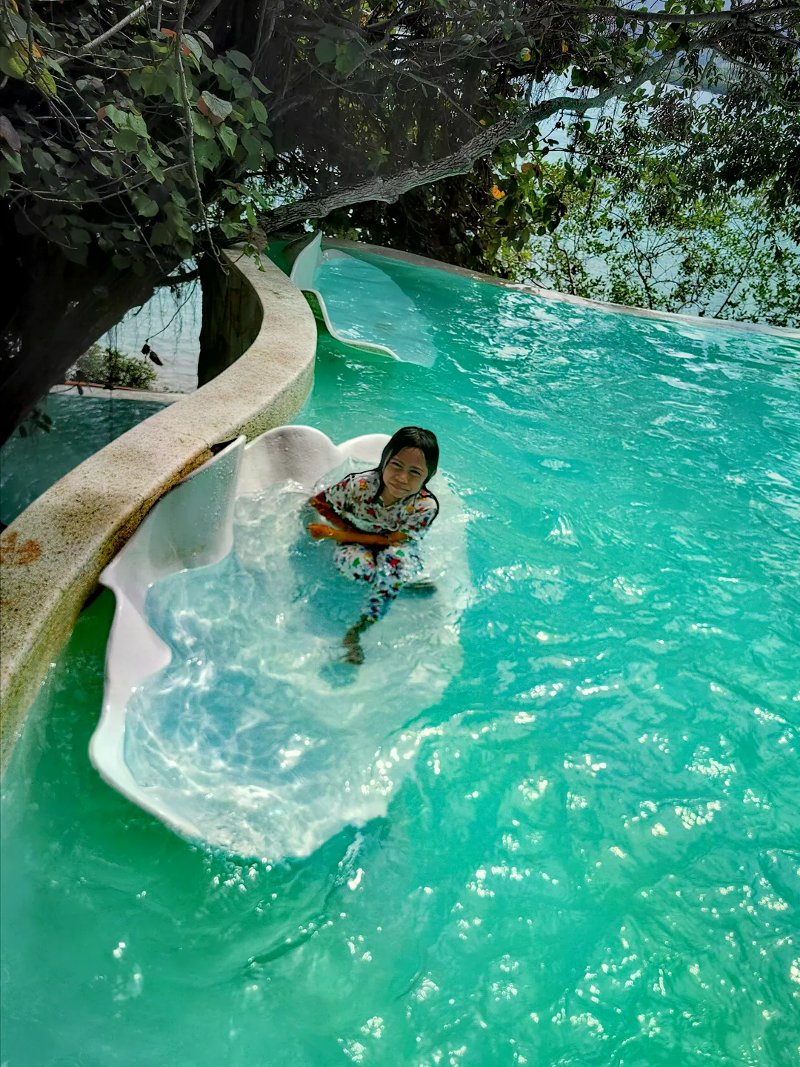 Girl smiling in a shell-shaped lounger in a turquoise infinity pool under hanging trees.