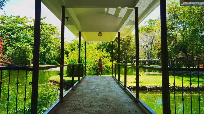 Child on a leaf-imprinted covered walkway overlooking a green lagoon and thatched pavilions.