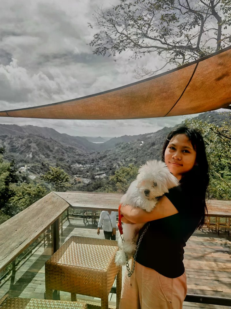 Woman holding a fluffy white dog on a shaded wooden deck overlooking green mountain ridges.