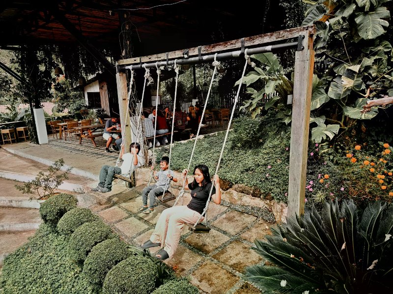 Three people on wooden rope swings beside a shaded garden dining patio at Charlie's Cup.