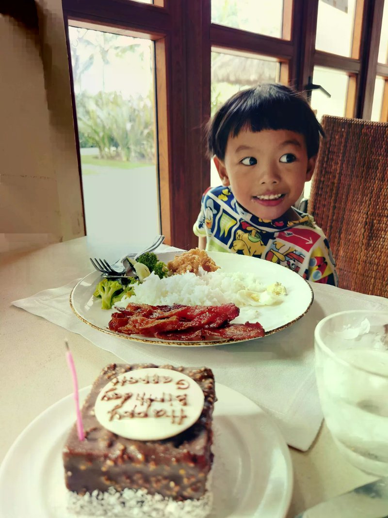 Smiling child at a resort table with Filipino breakfast and a lit Happy Birthday chocolate cake.