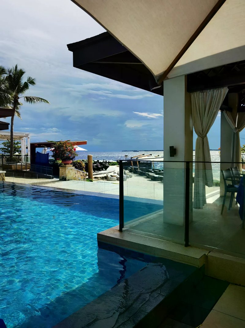 Infinity pool meeting the blue ocean beside a white-curtained pavilion and palm trees.