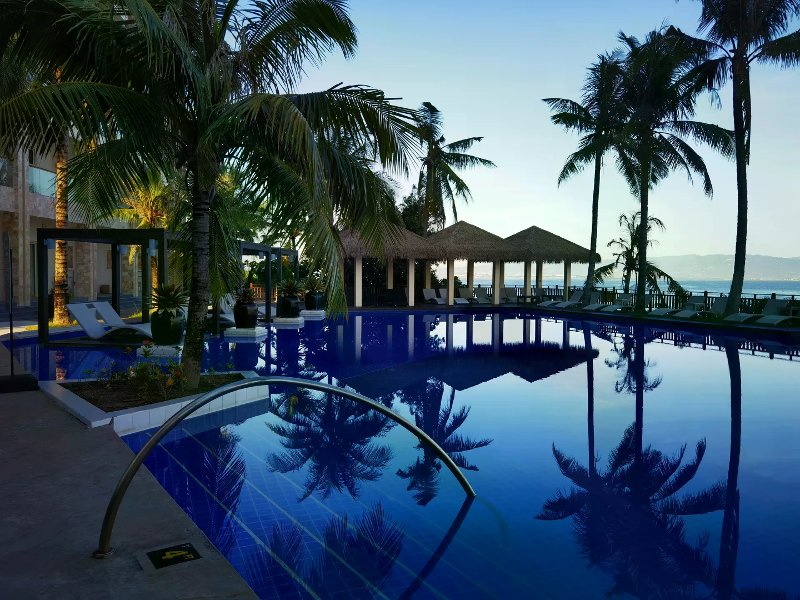 Calm infinity pool reflecting palms and thatched gazebos with ocean and mountains beyond.