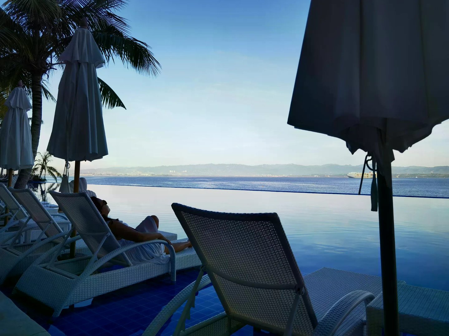 White wicker loungers along an infinity pool overlooking calm ocean and distant mountains.
