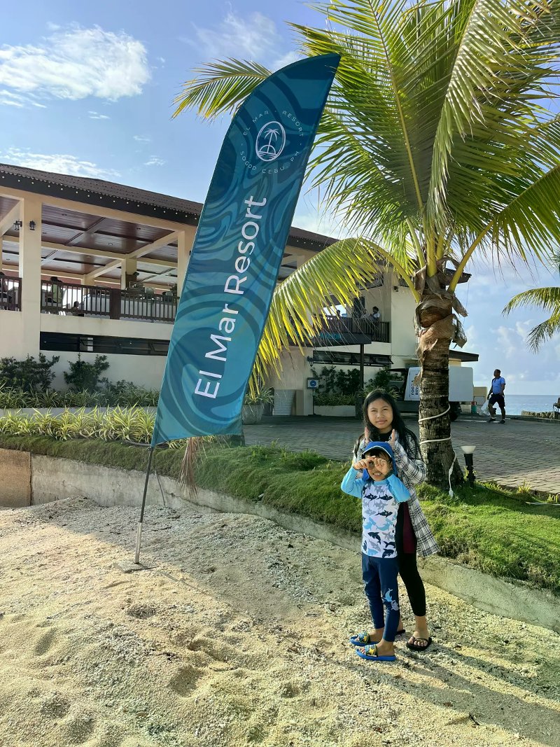 Guests beside a blue El Mar Resort banner with the beachfront building and palm behind them.