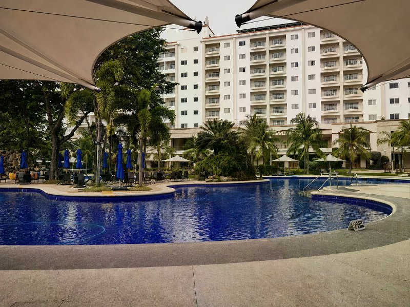 Large blue swimming pool with palms and a tall white resort hotel under an overcast sky.