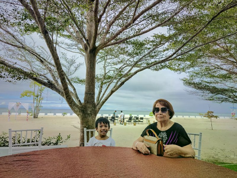 Elderly woman and boy at a shaded outdoor table overlooking a wide white-sand beach.