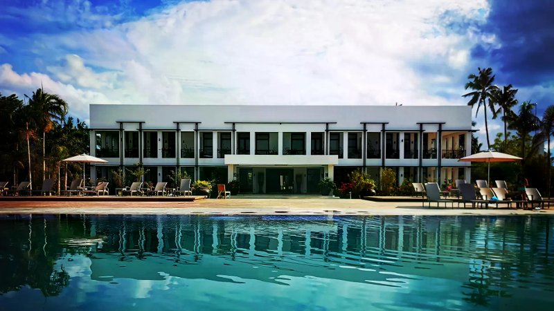 Modern two-story white resort building reflected in a large pool with lounge chairs and umbrellas.