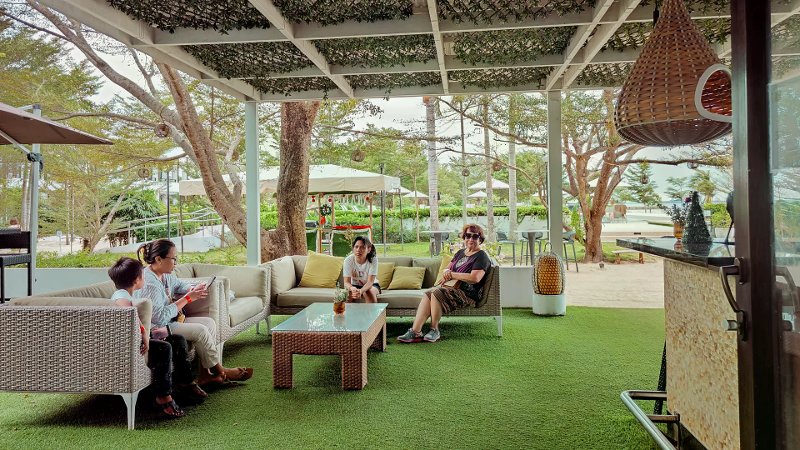 Guests on grey wicker sofas under a white pergola with artificial turf near a beach bar.