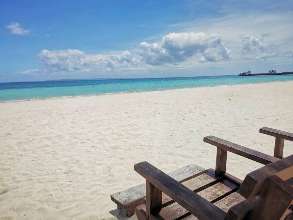 Wooden beach loungers sitting on the white sand beach of Anika Island Resort in Bantayan, facing clear turquoise ocean waters under a sunny blue sky with soft white clouds.