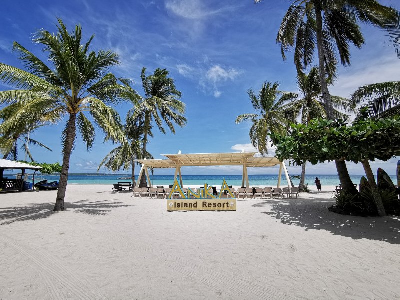세부 반타얀 섬 산타페에 있는 Anika Island Resort의 해변 전경. 야자수와 백사장, 푸른 하늘 아래 세워진 나무 리조트 표지판이 보인다.