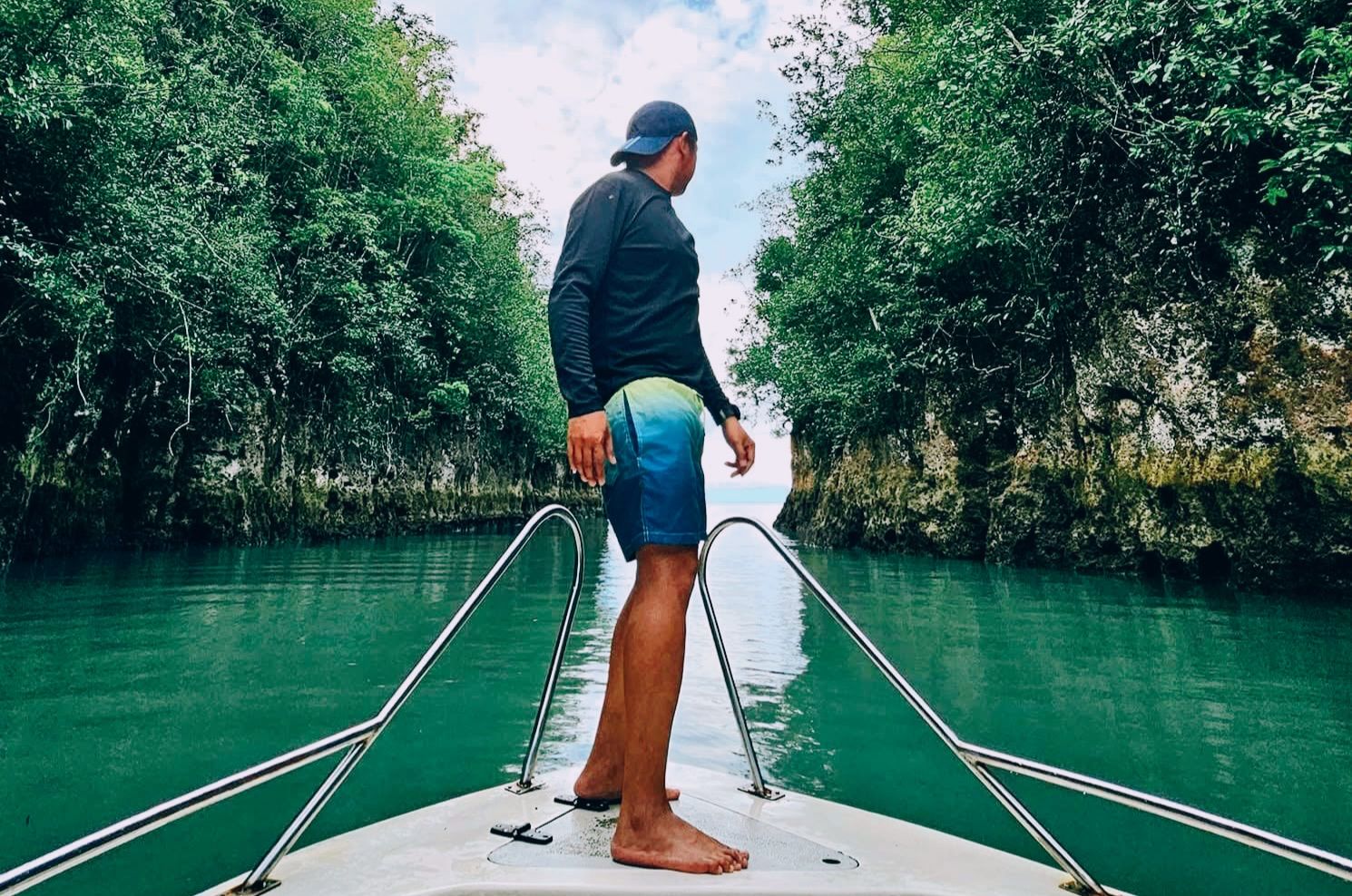 A man in a black long-sleeve rashguard, blue and yellow swim trunks, and a baseball cap stands on the bow of a boat facing away from the camera, navigating through the calm, green water of Bojo River lined with lush rock cliffs under a cloudy sky.
