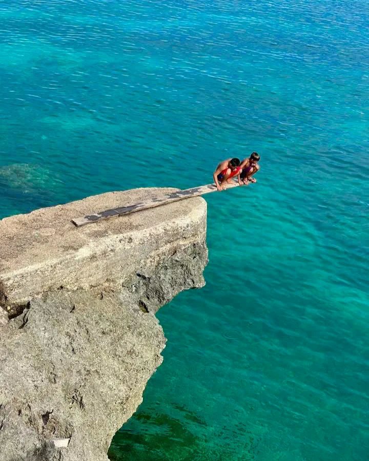 Two people preparing to jump from a wooden diving board built on a rocky cliff over clear turquoise ocean water at Buho Rock Resort