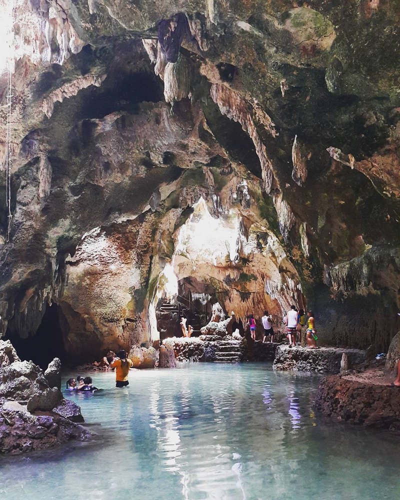 A large, cavernous opening inside Bukilat Cave in Camotes, showing natural limestone rock formations and stalactites hanging from the ceiling. Bright natural sunlight streams down from an opening above onto a group of visitors standing near a clear pool of water at the bottom.
