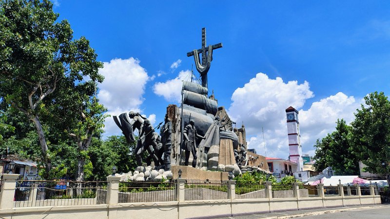 A wide shot of the Cebu Heritage Monument in Parian, Cebu City, featuring dark bronze sculptures depicting historical figures, a Spanish galleon, and a large cross under a bright blue sky with fluffy white clouds. A tall clock tower stands in the background to the right.