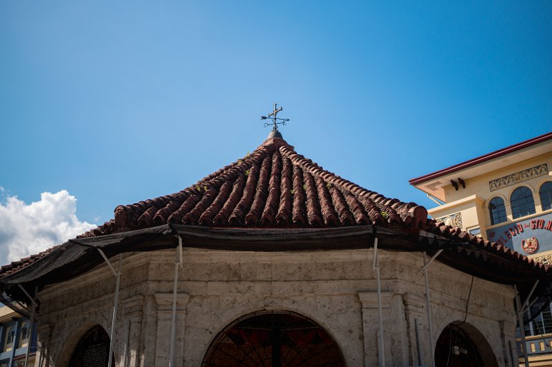 The exterior of the Magellan's Cross pavilion in Cebu, featuring an octagonal stone structure with arched openings and a red-tiled roof under a clear blue sky.