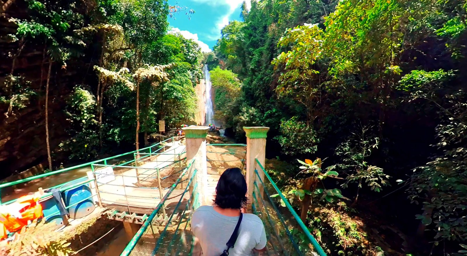 A view from behind a person on a metal footbridge with green handrails, looking toward the main towering waterfall of Mantayupan Falls in Cebu, surrounded by lush green foliage and bright sunlight.