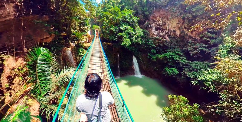 High-angle view from behind a person walking across a long wooden suspension bridge with blue railings, looking over a natural pool and small waterfall surrounded by dense tropical trees at Mantayupan Falls, Cebu.