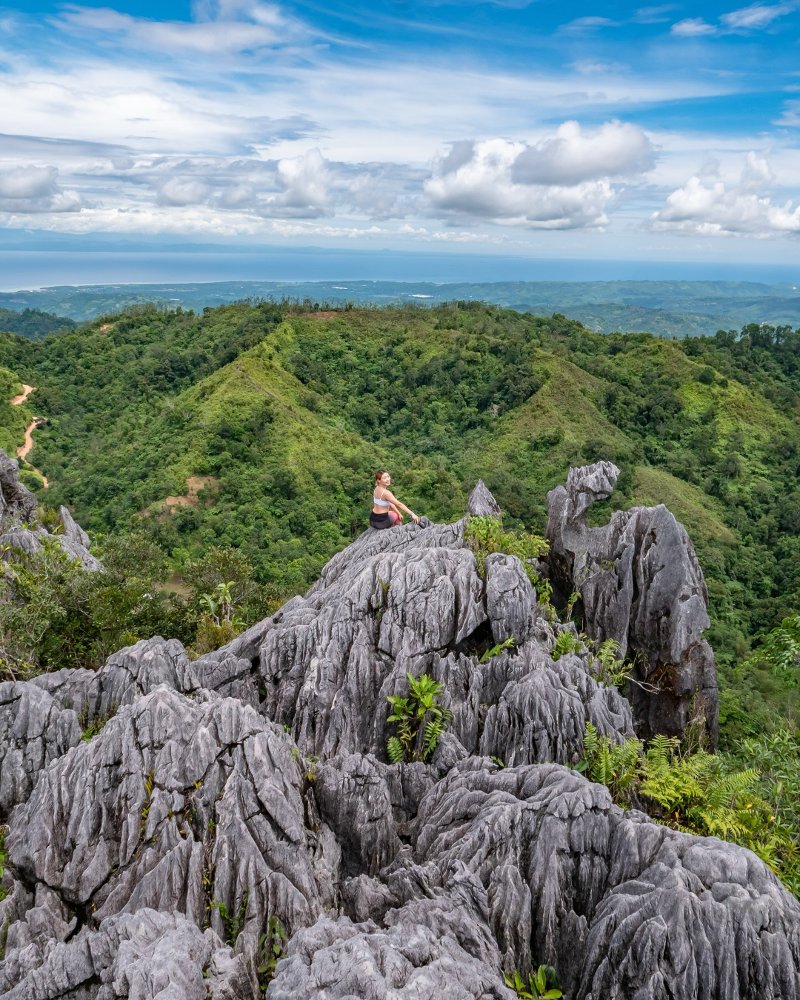 Wide landscape view of a female hiker sitting on a rugged limestone cliff at Mt. Mauyog, Cebu, surrounded by rolling green hills, distant coastline, and a cloudy blue sky.
