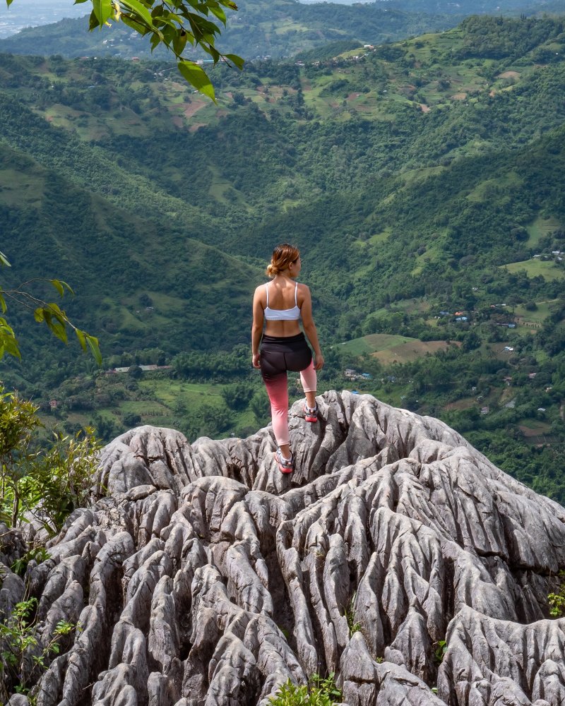 A woman in athletic wear standing on the rocky, brain-like limestone peak of Mt. Mauyog in Cebu, looking out over lush green mountain valleys.