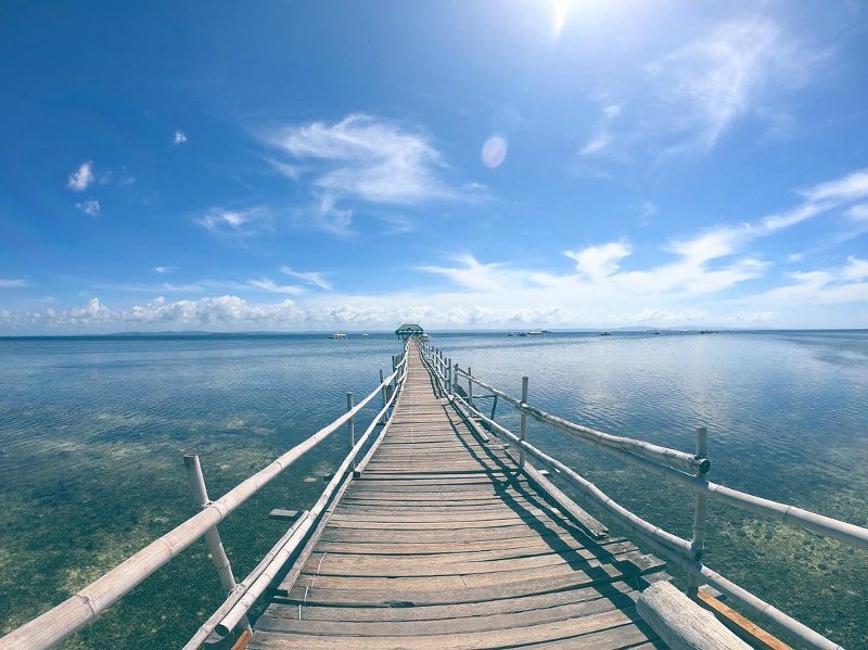 Long wooden boardwalk leading across clear shallow turquoise water toward Nalusuan Island under a bright blue sunny sky.