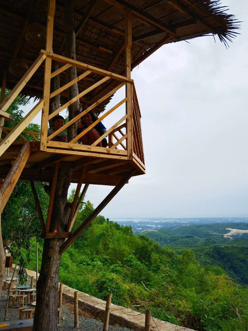 A wooden treehouse viewing deck built around a tree trunk at Nanda Cafe, overlooking lush green hills and a distant landscape under an overcast sky.