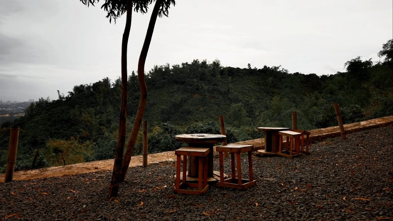 An outdoor seating area at Nanda Cafe featuring rustic wooden tables made from large spools and matching stools, set on a gravel surface with a backdrop of a dense, forested hill.