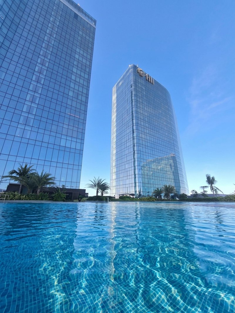 The outdoor swimming pool at NUSTAR Resort Cebu surrounded by palm trees, reflecting the modern glass towers under a clear blue sky.