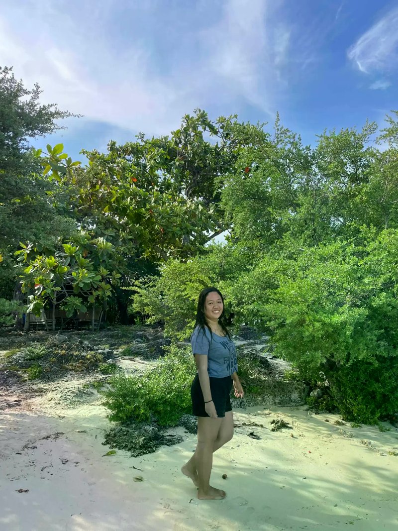 A woman standing outdoors in front of dense, lush green trees and coastal vegetation under a bright blue sky