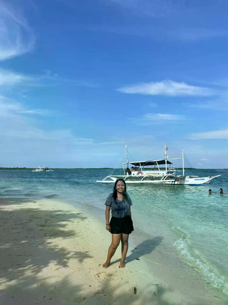 A woman smiling on a sandy beach on Olango Island with clear blue water, a outrigger boat moored near the shore, and a partly cloudy blue sky in the background.