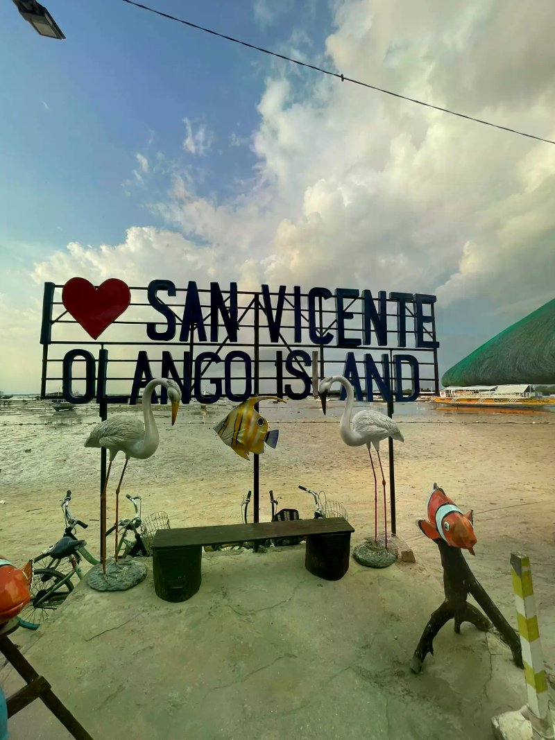 A metal welcome sign reading "I ❤️ SAN VICENTE OLANGO ISLAND" decorated with fish and flamingo sculptures on a coastal shoreline under a cloudy sky