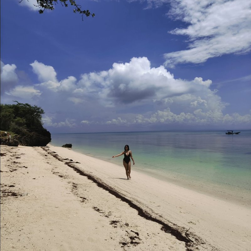 A person in a black swimsuit walks along the white sand shoreline at Paradise Beach in Bantayan Island, Cebu, surrounded by clear turquoise water, rocky cliffs, and a bright blue sky with fluffy white clouds.