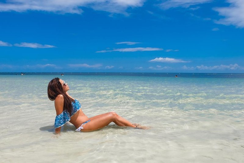 A woman in a blue pattern bikini sunbathing in shallow clear water at Santiago Bay Beach in Camotes Islands under a clear blue sky.