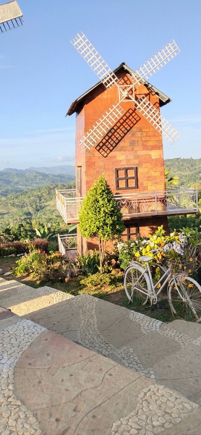 A decorative white bicycle parked in a garden area in front of a brick windmill structure, set against a vast backdrop of lush green mountainous landscapes at Sirao Garden, Cebu.