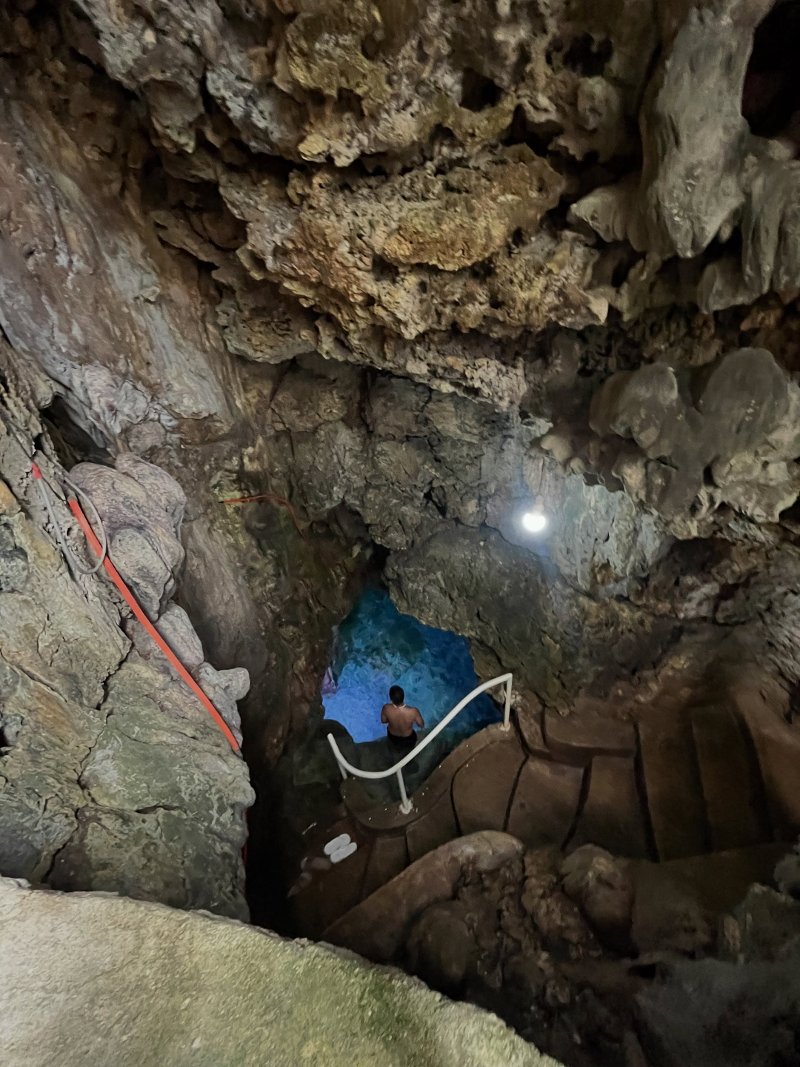 Looking down from a steep concrete staircase with white safety handrails into a subterranean limestone cavern leading to a glowing, turquoise freshwater pool at the bottom of Timubo Cave.
