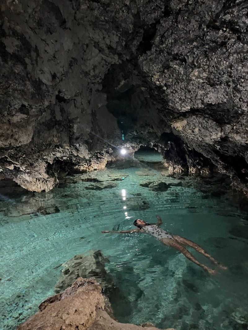 A woman wearing a swimsuit floats peacefully on her back in the crystal-clear freshwater pool inside Timubo Cave, surrounded by rough limestone cavern walls under gentle underground ceiling lights.