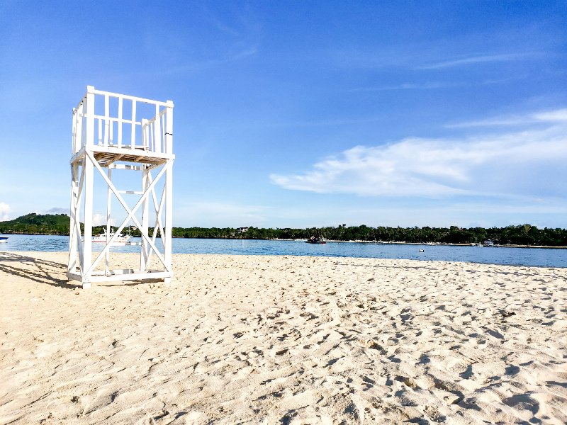 A white wooden lifeguard tower on a wide white sand beach with calm blue water and a distant green shoreline under a bright blue sky.