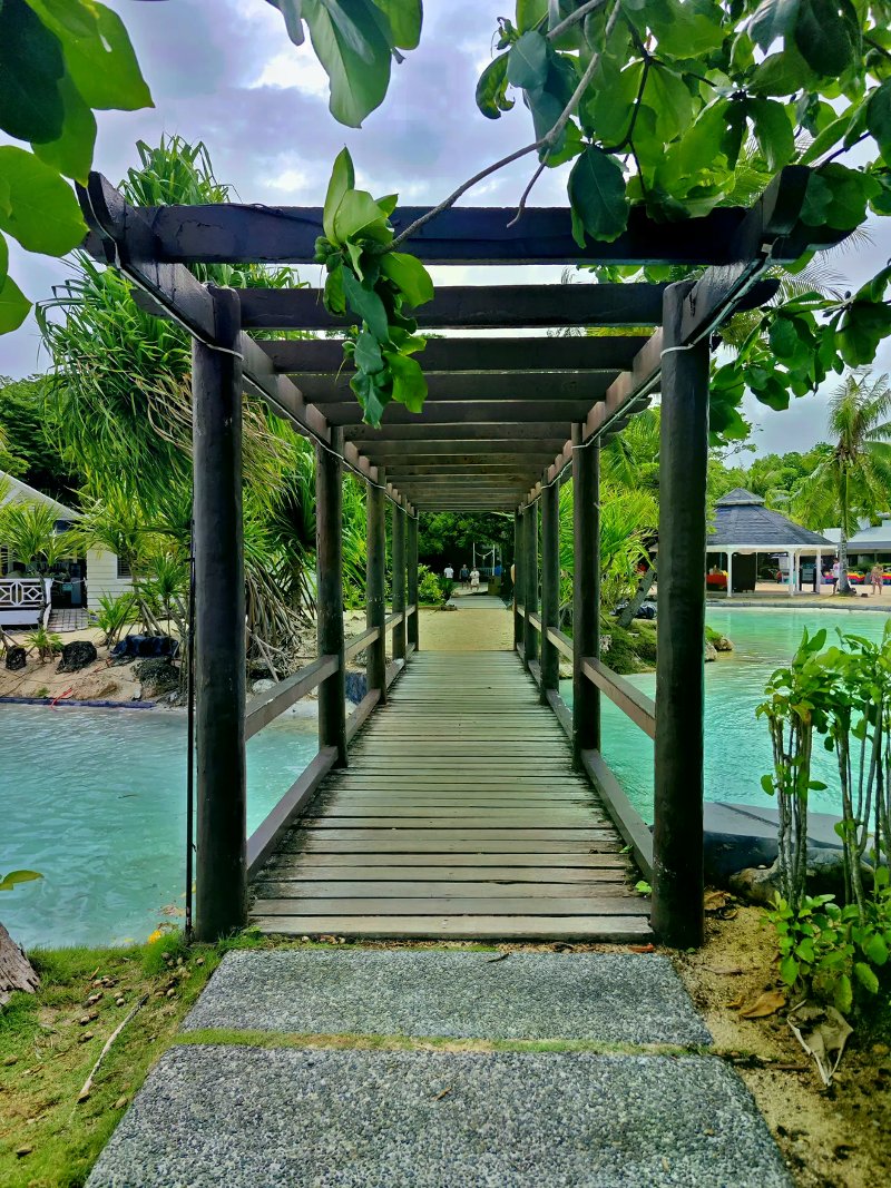 Wooden walkway under a dark pergola crossing a bright turquoise lagoon toward resort gardens.