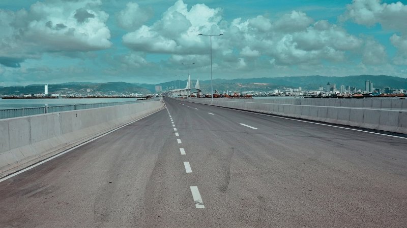 An empty multi-lane paved bridge stretching toward distant mountains and a city skyline under a cloudy blue sky.