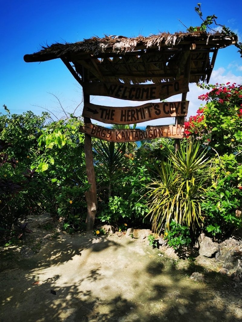Wooden welcome sign for Hermit's Cove in Aloguinsan, surrounded by vibrant green tropical foliage and red flowers.