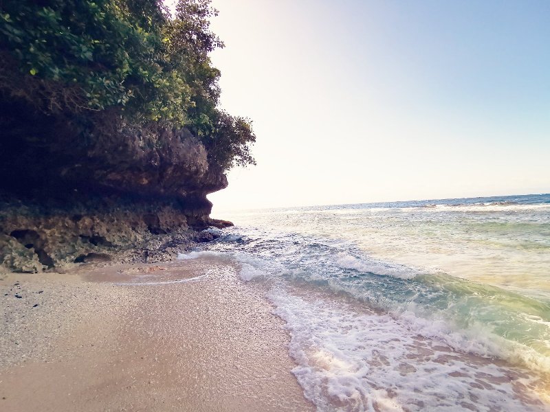 Shoreline view at Hermit's Cove in Aloguinsan showing gentle waves rolling onto the sand beside a rocky cliff overhang.