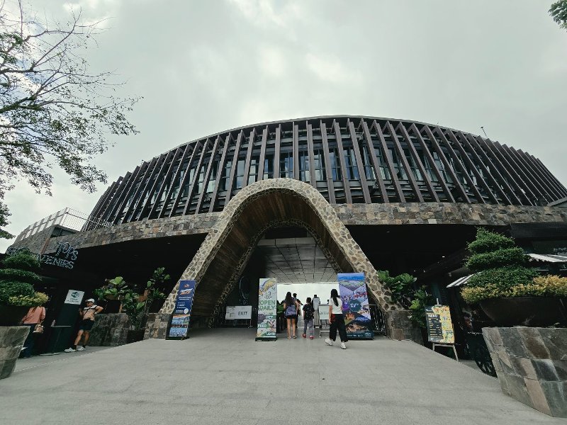 Main entrance of the modern circular Tops Cebu structure featuring a large triangular stone archway and slatted wooden facade under a cloudy sky.