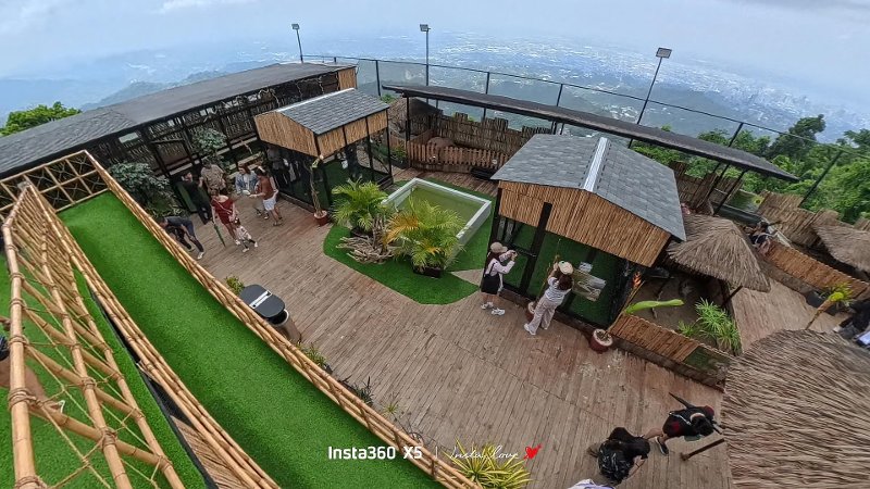 High-angle wide shot of the outdoor Tops Petting Park featuring wooden deck pathways, animal shelters, green turf, and scenic mountain views.