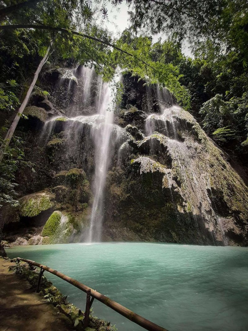 Mid-shot of Tumalog Falls in Oslob, featuring mist-like water cascading down rock formations into a vibrant turquoise natural pool below.